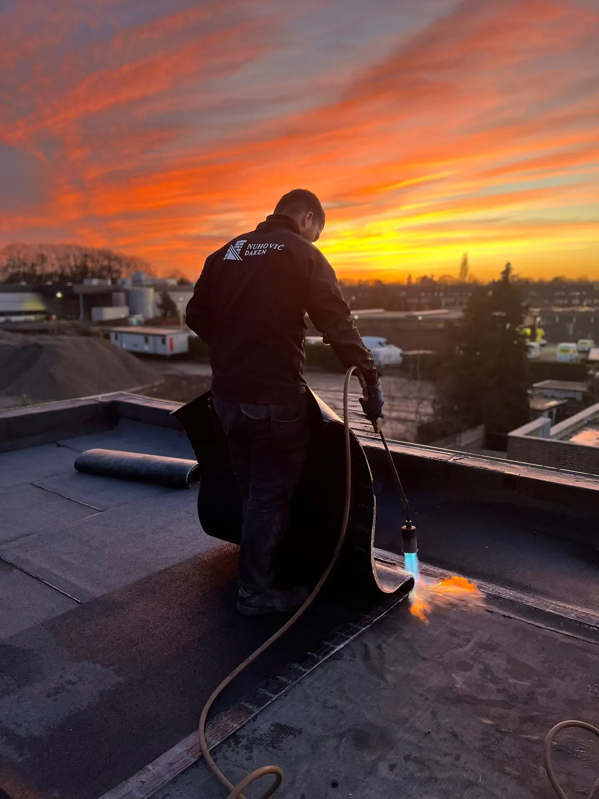 Dakdekker van Nuhovic Daken brandt bitumen dakbedekking op een plat dak bij zonsondergang